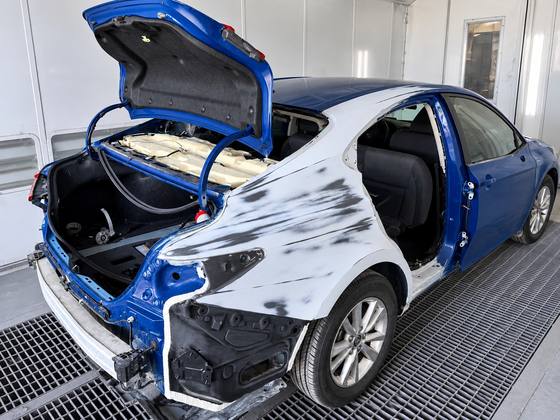 The same Camry mid-repair inside the paint booth, rear quarter stripped to guide coat with the trunk lid and bumper removed